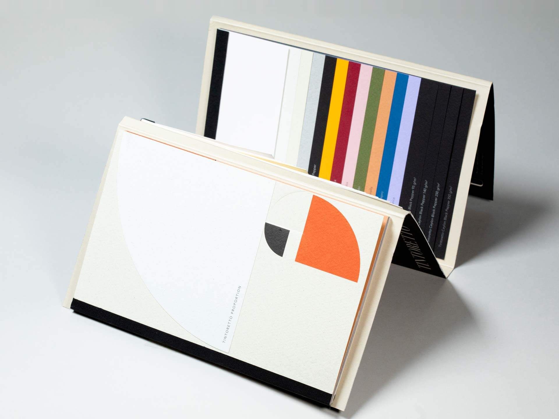 Due libretti campioni rilegati aperti a tenda su superficie grigia, che mostrano un ventaglio di strisce di carta colorate in una gamma completa di colori e una copertina con design geometrico astratto arancione e grigio. I libretti di presentazione e campionario personalizzati aiutano imprenditori e aziende a presentare materiali, finiture e opzioni di brand in un formato stampato premium.