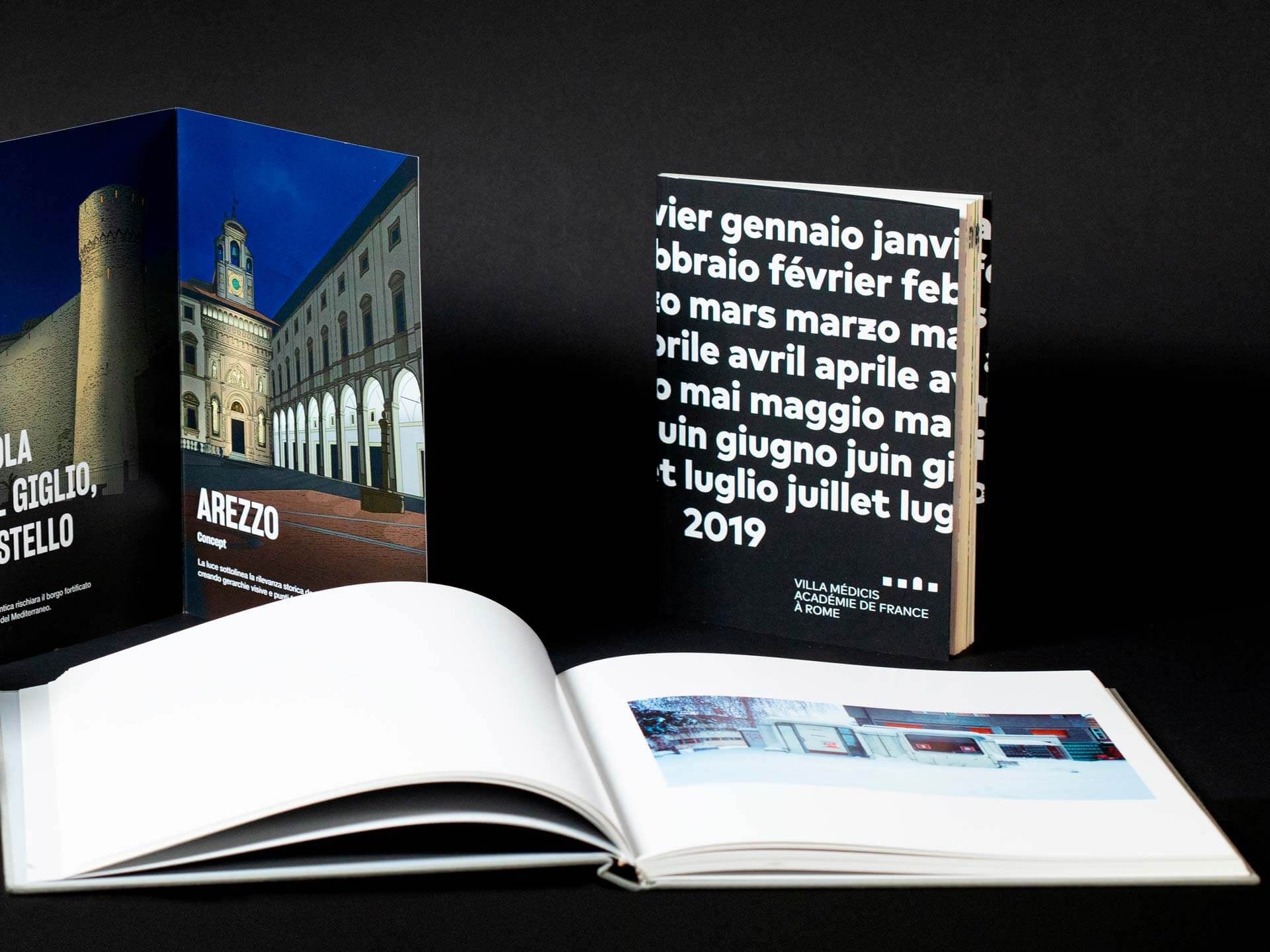 Due libri con copertina rigida personalizzati aperti su sfondo scuro — un libro fotografico architettonico su Arezzo con fotografia notturna di torri storiche, e uno con copertina tipografica calendario multilingue 2019. Stampa di libri personalizzati premium per imprenditori e aziende — libri fotografici rilegati, pubblicazioni brandizzate e cataloghi aziendali con finiture di qualità.