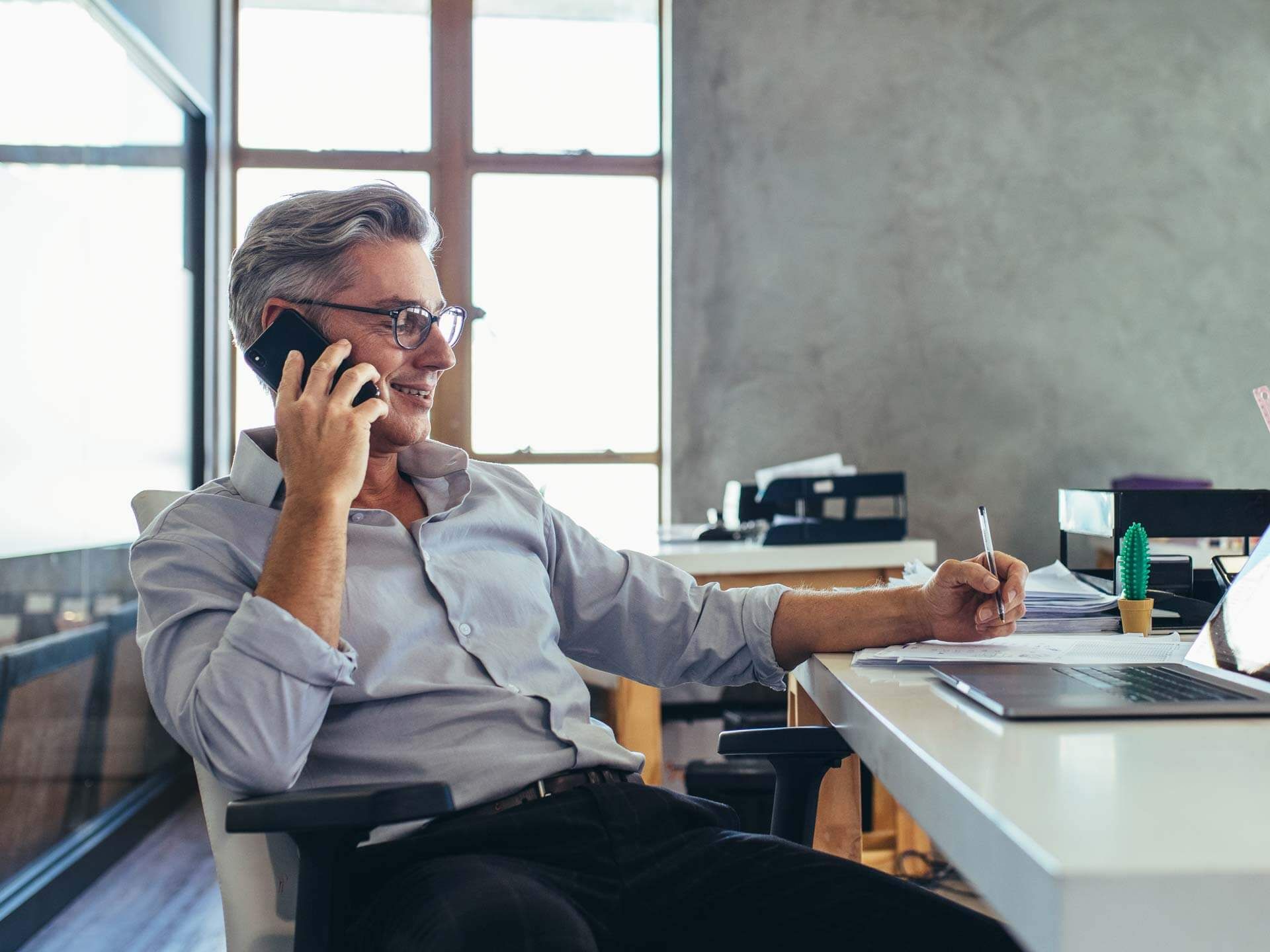 Un imprenditore maturo con occhiali che parla al telefono mentre prende appunti alla scrivania di un ufficio moderno, con luce naturale da una grande finestra sullo sfondo. I servizi di stampa professionale per imprenditori — dai biglietti da visita alle intestate, dai cataloghi alla cancelleria brandizzata — aiutano le aziende a comunicare in modo professionale ed efficiente.