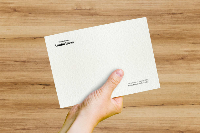 Una mano regge una busta bianca Tintoretto Neve con superficie finemente testurizzata e logo in corsivo su fondo legno. La carta Tintoretto Neve è nota per la sua texture vergata e il tono bianco brillante. Le buste personalizzate in Tintoretto Neve sono ideali per corrispondenza di pregio e cartoleria premium.
