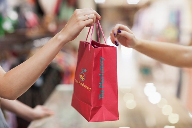 Due mani si scambiano una vivace shopper in carta rossa con logo personalizzato stampato in un contesto retail. La stampa di alta qualità su questa shopper rinforza la visibilità del brand al punto vendita. Le buste shopper in carta personalizzate sono uno strumento di marketing efficace per negozi e boutique.
